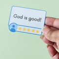 A hand holding a sticker with the text 'God is good!' surrounded by a blue frame and five yellow stars on a yellow background.