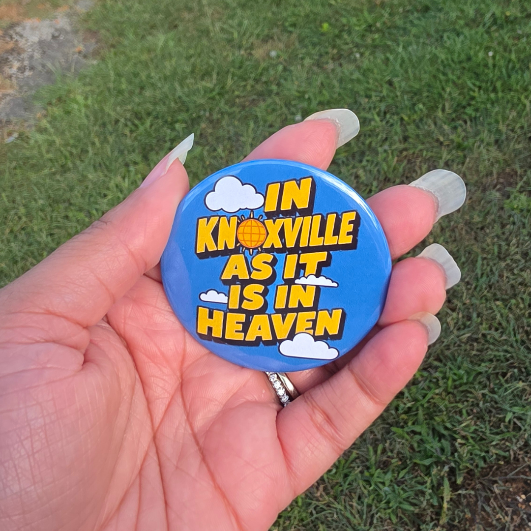 A hand with long nude manicured nails holds a round blue button badge outdoors on a grassy lawn. The badge features the text 'IN KNOXVILLE AS IT IS IN HEAVEN' in yellow letters with white clouds and a small sun graphic.