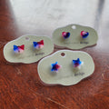 Three pairs of resin stud earrings in red, white, and blue colors, displayed on green earring cards with 'Nug & Carol Design' written on them.