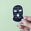A hand holding a black vinyl sticker with white text and design elements resembling a mask.