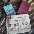 A beige 'THE BIBLE BAG' tote bag with black text, displayed alongside an Bible, a notebook, and pens on a textured background.