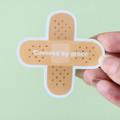 A hand holding a beige-colored sticker with the phrase 'Covered by grace' written in a central position, designed to resemble a bandage.