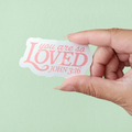 A hand holding a white sticker with the text 'you are so LOVED' in pink letters, and the Bible reference 'John 3:16' in smaller text.