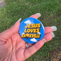 A hand holding a round blue button badge with the text 'JESUS LOVES KNOXVILLE' in yellow and white clouds, photographed outdoors on grass.
