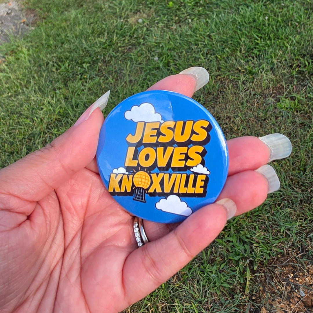 A hand holding a round blue button badge with the text 'JESUS LOVES KNOXVILLE' in yellow and white clouds, photographed outdoors on grass.