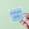 A hand holding a blue square sticker with the text 'Read your Bible' written in a cursive black font.