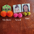 A set of three pairs of fluffy pom pom earrings in orange, pink, and green colors, displayed on a wooden surface with tags indicating the sizes as Mini, Small, and Medium.